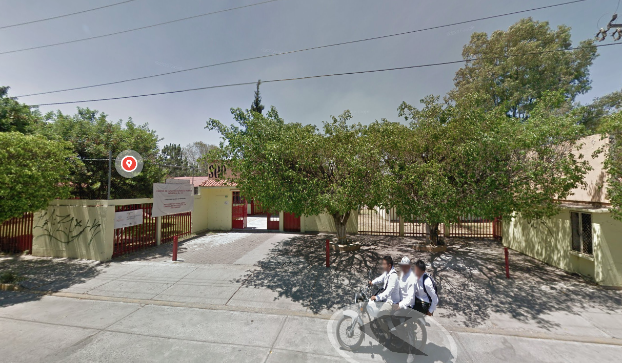 Fotografía en la que se muestra una explanada. Al fondo de la explanada se encuentra la entrada del plantel, conformada por muros verdes intercalados con rejas rojas. Una de las rejas del lado izquierdo es la que da acceso y sobre ell<a hay una lona con los datos del plantel.